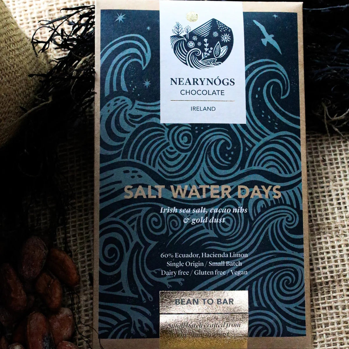 A packaged chocolate bar from Nearynógs Chocolate, labeled 'Salt Water Days', displayed on a burlap surface with seaweed and cocoa beans as styled props.