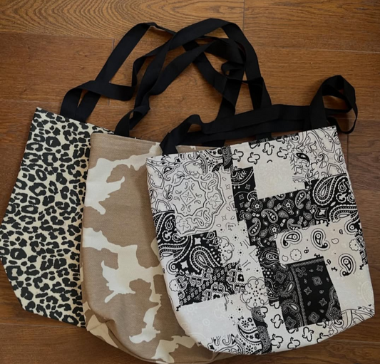 Three repurposed fabric tote bags with black handles, displayed on a wooden floor. The bags feature different prints: leopard print, tan and beige camouflage, and a black-and-white patchwork bandana pattern.