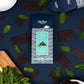 A package of Skellugs Chocolate Mint Dark Chocolate Shards is displayed in a styled flat-lay on a dark navy blue background, surrounded by broken chocolate pieces and fresh mint leaves.