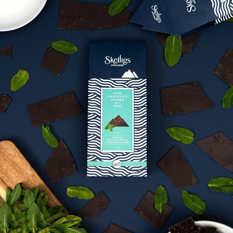 A package of Skellugs Chocolate Mint Dark Chocolate Shards is displayed in a styled flat-lay on a dark navy blue background, surrounded by broken chocolate pieces and fresh mint leaves.