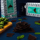A styled product shot of dark chocolate pieces stacked in the center, with mint leaves and branded packaging boxes in the background on a dark blue surface.